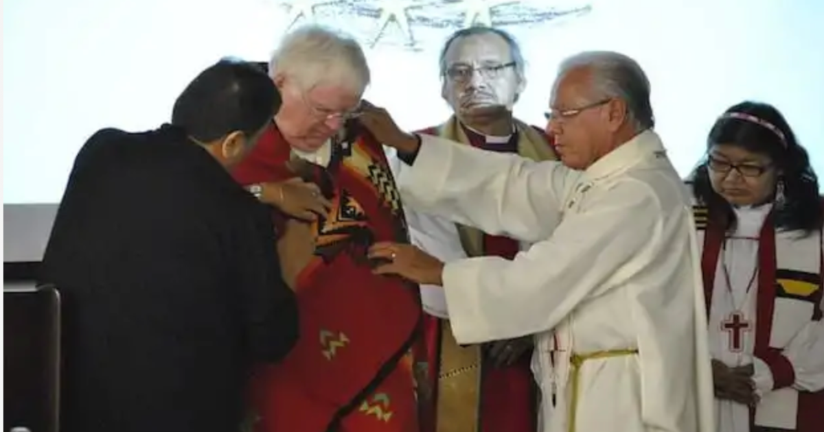 Indigenous bishops, clergy and Elders wrapped a sunset red Pendleton blanket over Archbishop Michael Peers and prayed over him, in tribute to the Archbishop's apology paving the way for healing and reconciliation. The apology was delivered by then-Primate Michael Peers to the National Native Convocation in Minaki, Ontario. Pictured with the Archbishop are former indigenous ministries co-ordinators Donna Bomberry and the Rev. Canon Laverne Jacobs as well as Bishops Adam Halkett and Lydia Mamakwa looking on